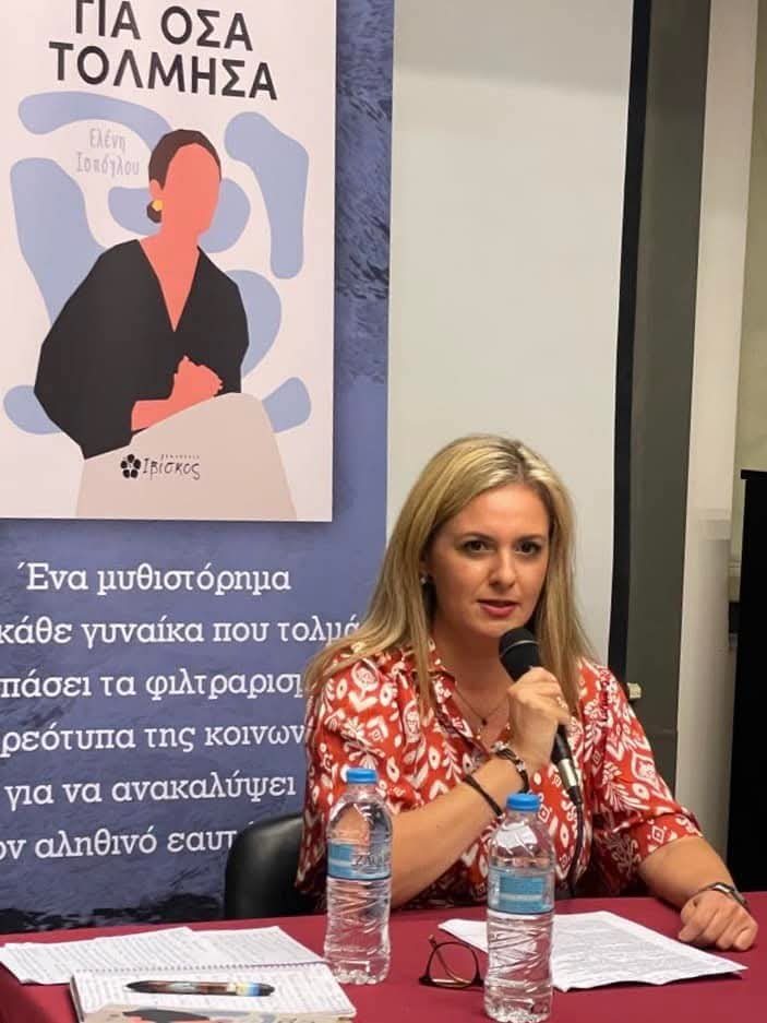 Η Ελένη Ισπόγλου μιλά στο μικρόφωνο μπροστά στο banner του βιβλίου.
